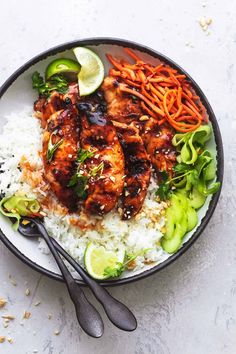 a bowl filled with rice, carrots and chicken next to two serving utensils