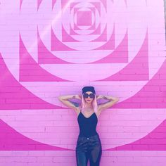 a woman standing in front of a pink wall with her arms behind her head and sunglasses on