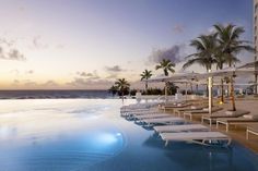 an empty swimming pool with lounge chairs and umbrellas next to the ocean at sunset