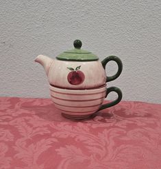 a ceramic teapot with an apple painted on the front and sides, sitting on a pink tablecloth