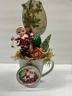 an elf figurine sitting in a coffee cup filled with christmas decorations and ornaments