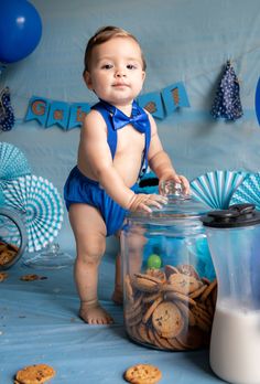 a baby in a blue outfit standing next to a jar full of cookies and milk