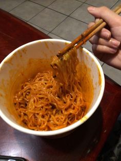 a person holding chopsticks over a bowl of noodles with sauce on the table