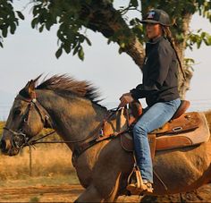 a woman riding on the back of a brown horse next to a tree and dirt road