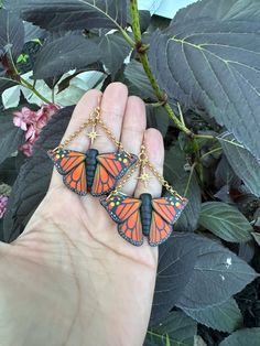 a hand holding two orange and black butterfly shaped earrings on it's fingers, with leaves in the background