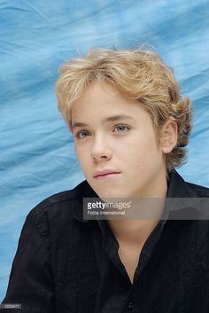 a young man with blonde hair sitting in front of a blue background wearing a black shirt