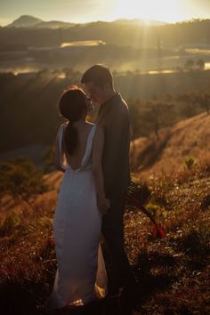 a bride and groom standing on top of a hill at sunset with the sun behind them