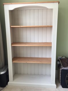 an empty white bookcase with wooden shelves in the corner next to a black suitcase