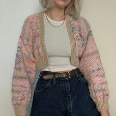 a woman with blonde hair wearing a pink cardigan and jeans, standing in front of a white wall