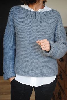 a woman standing in front of a wooden dresser wearing a blue sweater and black pants