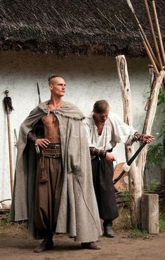 two men dressed in medieval clothing standing next to each other near a white building with thatched roof