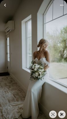 a woman in a wedding dress standing by a window holding a bouquet and looking at the camera