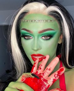 a woman with white hair and green makeup is holding her hand up to her face