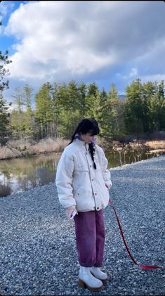 a woman in white jacket holding a red leash on gravel ground next to water and trees