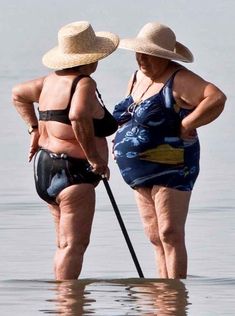 two women in swimsuits and hats stand on the beach with their backs to each other