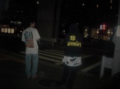 two people standing on the street at night, one is wearing a hoodie and the other has a t - shirt