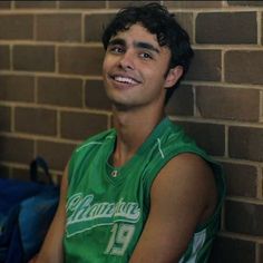 a young man sitting against a brick wall wearing a green jersey and smiling at the camera