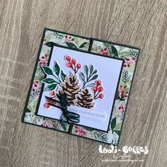 a christmas card with pine cones and holly berries on it, sitting on a wooden surface