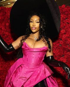 a woman in a pink dress and black hat standing next to red rose bushes with her arms out