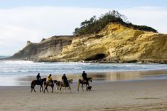 four people riding horses on the beach near an ocean and rock formation in the background