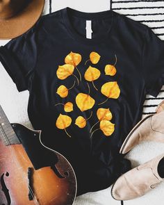 T-shirt featuring a botanical illustration of golden yellow Aspen tree leaves in the fall. Fall in love with our perfect autumn graphic tee you won't find anywhere else. Printed on a Black shirt, made from durable lightweight cotton.  Unisex fit - boxy and long. T-SHIRT SIZING: *All measurements in inches XSLength 27 / Width 16 ½ SLength 28 / Width 18 MLength 29 / Width 20 LLength 30 / Width 22 XLLength 31 / Width 24 2XLLength 32 / Width 26 3XLLength 33 / Width 28 How it's made: * All prints are Yellow Graphic Tee For Fall, Casual Yellow T-shirt For Fall, Yellow Cotton T-shirt For Fall, Mustard Short Sleeve Tops For Fall, Yellow T-shirt With Screen Print For Fall, Black T-shirt With Floral Print For Fall, Casual Fall T-shirt With Plant Print, Casual T-shirt With Plants Print For Fall, Casual Plants Print T-shirt For Fall