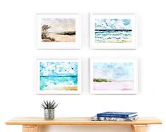 four watercolor paintings hang on the wall above a wooden table with a plant and books