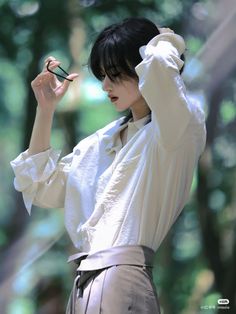 a woman with black hair and white shirt is holding scissors up to her head while standing in front of trees