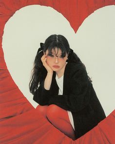 a woman sitting on top of a red heart shaped pillow with her hands to her face