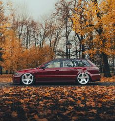 a red car parked in front of some trees with leaves on the ground and one person standing next to it