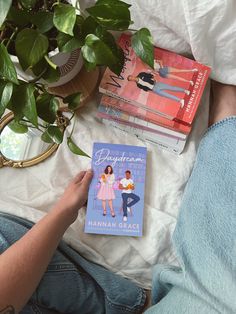 a person sitting on a bed reading a book next to a potted plant and other books