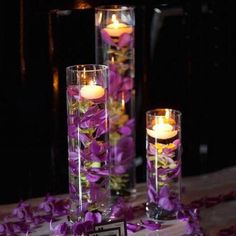 three tall clear vases filled with purple flowers and lit candles on a white table