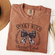 Embrace your spooky side with our "Spooky Witch Social Club" T-Shirt. Perfect for Halloween or any day you feel like unleashing your inner witch, this tee features a beautifully detailed graphic of a lace bow with the playful phrase "Witches Be Crazy." Ideal for fans of gothic and witchy aesthetics, this shirt is sure to be a conversation starter. Whether you're casting spells or just enjoying some Halloween fun, this shirt is a must-have addition to your wardrobe. Halloween Cotton T-shirt With Letter Print, Halloween Letter Print Cotton T-shirt, Spooky Pre-shrunk T-shirt For Fall, Fall Witchy Graphic Print T-shirt, Crew Neck Cotton T-shirt For Halloween, Spooky Custom Print Halloween T-shirt, Witchy Halloween Crew Neck T-shirt, Spooky Fall T-shirt Pre-shrunk, Spooky Long Sleeve T-shirt With Custom Print