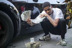 a man squatting next to a car holding money