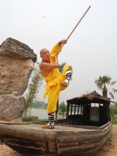 a man in yellow is practicing martial moves on a wooden boat at the riverbank