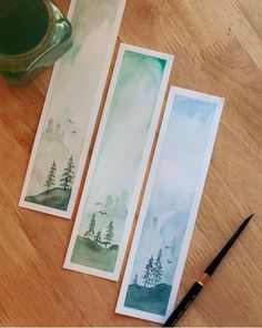 three watercolor bookmarks sitting on top of a wooden table next to a cup