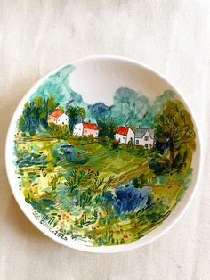 a small bowl with houses painted on the side and trees in the background, sitting on a white table cloth