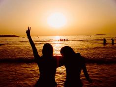 two women standing on the beach at sunset with their arms in the air and hands up