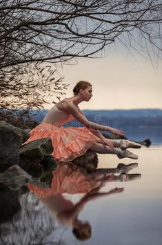 a woman in an orange dress is sitting on rocks by the water with her reflection