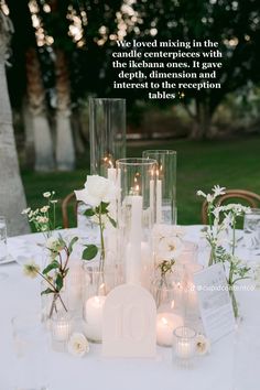 a table topped with candles and flowers on top of a white table cloth covered in glass vases