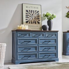 a blue dresser with flowers and candles on it in a white room next to a painting