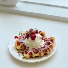 a waffle topped with whipped cream and raspberries on a white plate next to a window