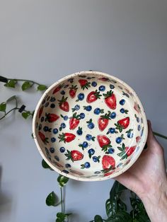 a hand holding a bowl with strawberries and blueberries painted on the inside, in front of a plant