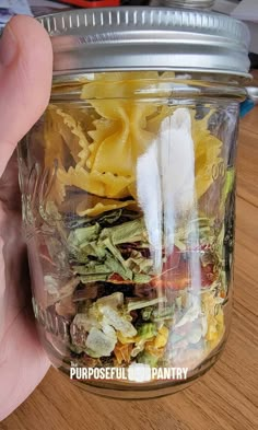 a person holding up a jar filled with pasta and other food items on top of a wooden table