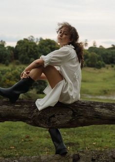 a woman sitting on top of a tree branch with her legs crossed and wearing rain boots