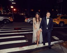 a man and woman walking across a cross walk at night in formal wear, with cars behind them