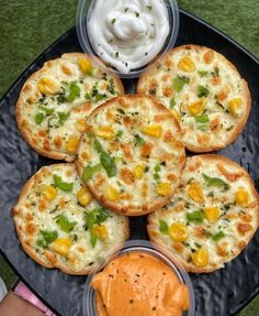 four small pizzas with different toppings on a black platter next to some dipping sauce