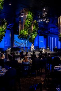 people are sitting at tables in front of a stage with plants hanging from the ceiling