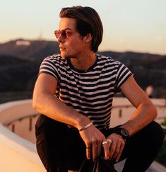 a man sitting on the edge of a wall with his hand on his hip while wearing sunglasses