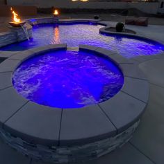 an outdoor hot tub lit up with blue lights in the evening time, surrounded by stone steps and fire pit