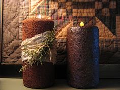 two lit candles sitting next to each other in front of a quilted wall hanging